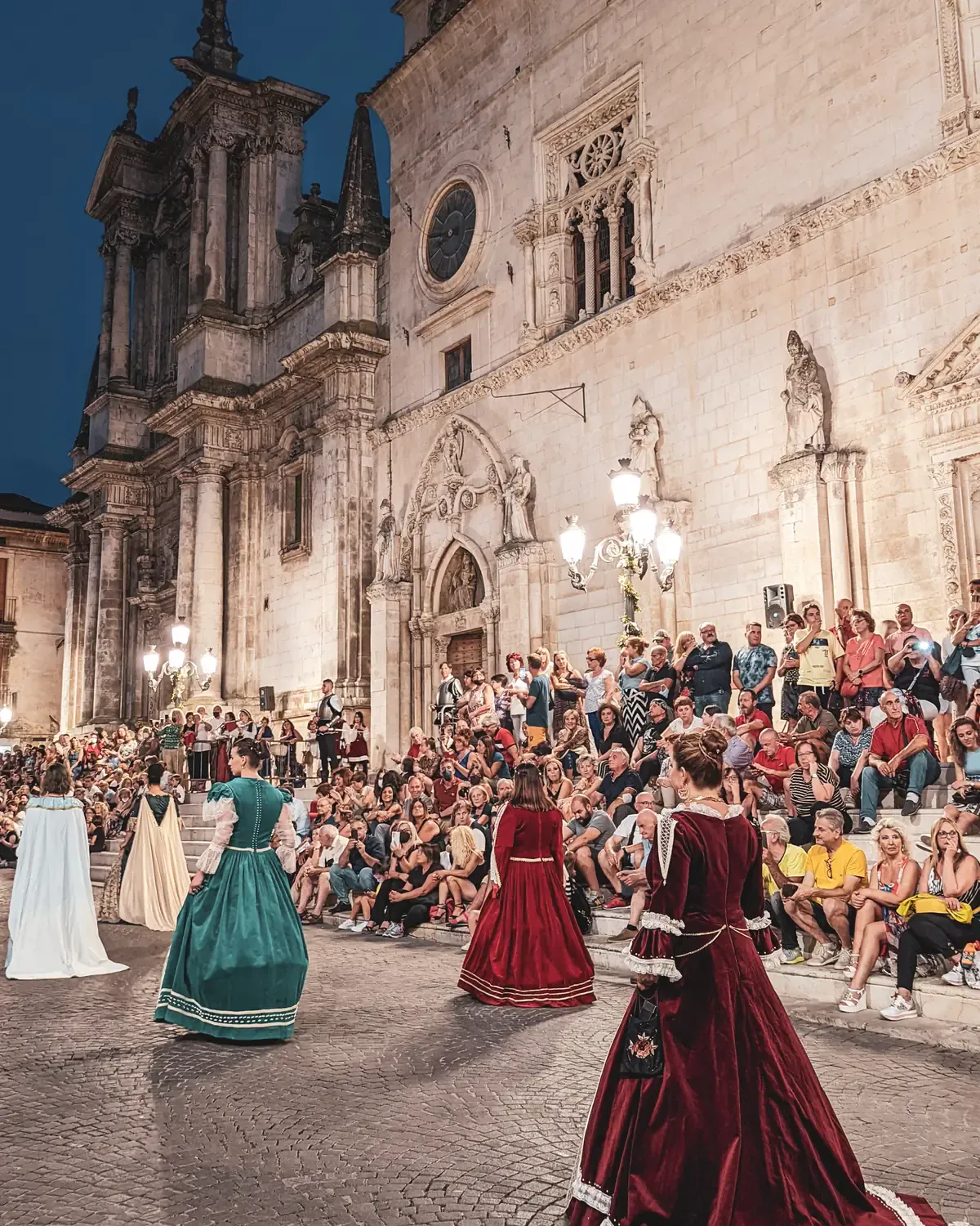 Frauen in historischen Kostümen tanzen abends vor dem Palazzo dell’Annunziata in Sulmona, auf den Stufen sitzt das Publikum